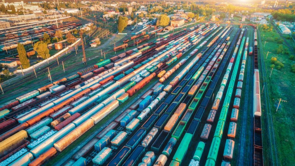 Flygfoto över godståg. Färgglada lastvagnar på järnvägsstationen. Vagnar med gods på järnväg. Tung industri. Industriellt landskap med tåg, järnvägsplattform vid solnedgången. Toppvy. Depå